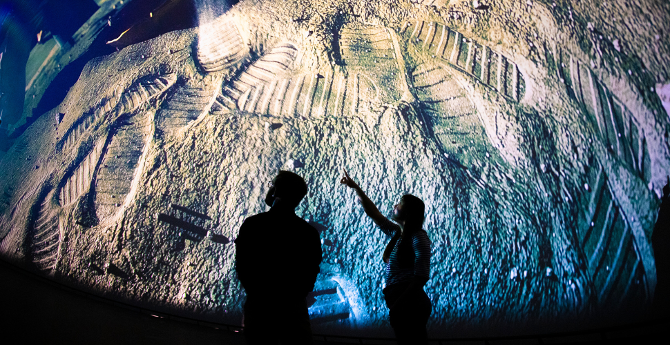 Two people stand silhouetted in front of a huge curved screen showing a close‑up of textured lunar-style ground covered in boot prints, as one person points up towards the display.